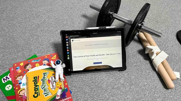 Tablet with FitFinanceDad open next to kids' learning books, dumbbell, and wrist roller