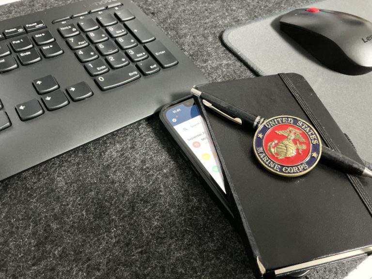 A black notebook with a United States Marine Corps emblem and pen on top, next to a smartphone showing the FitBudget app on a desk.