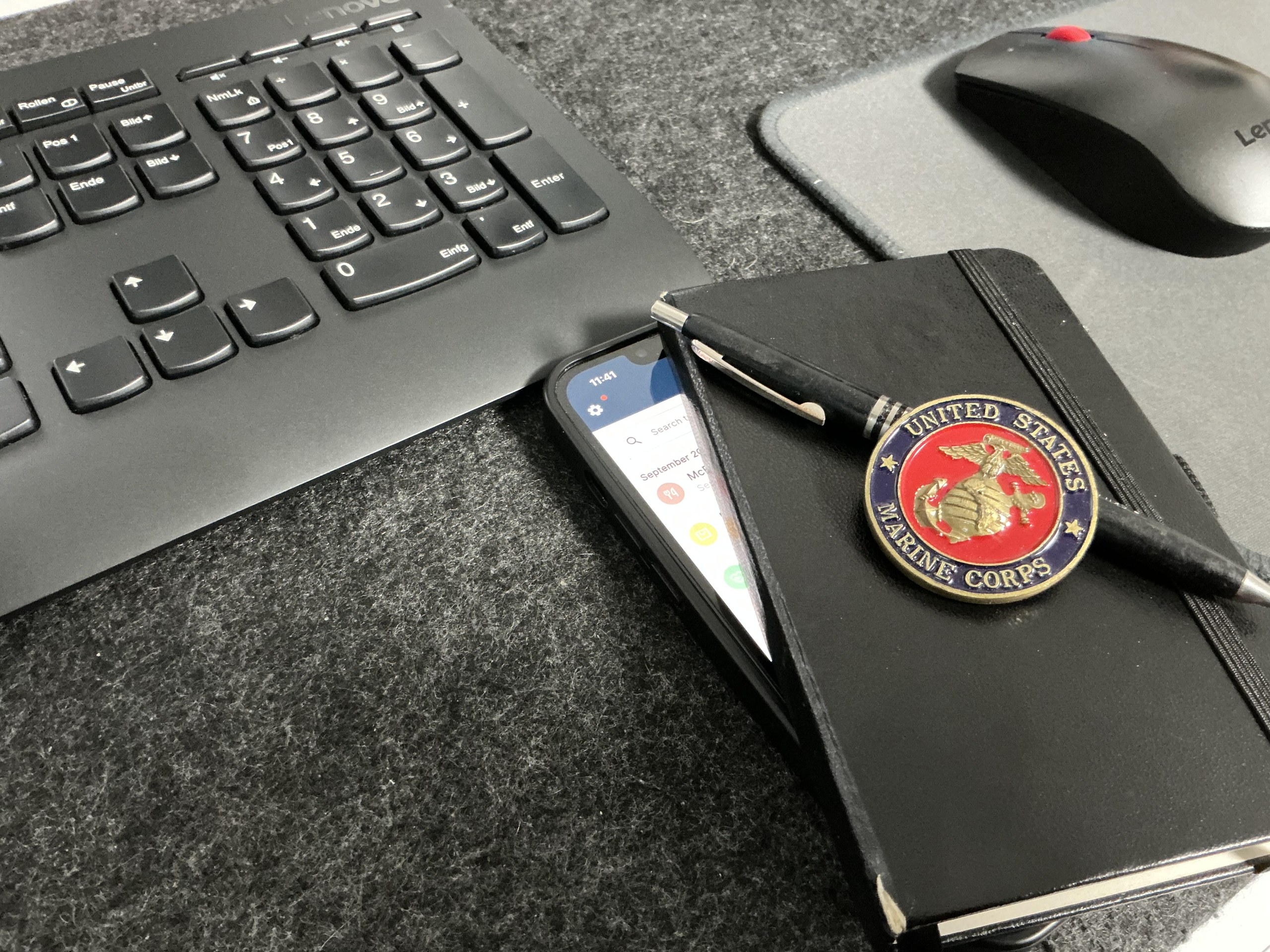 A black notebook with a United States Marine Corps emblem and pen on top, next to a smartphone showing the FitBudget app on a desk.